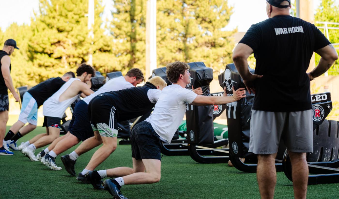 Linemen drive a blocking sled while a coach watches